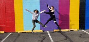 Bonnie Roseman and Trish Sunderland jumping energetically in a parking lot in front of a brightly painted striped brick wall.